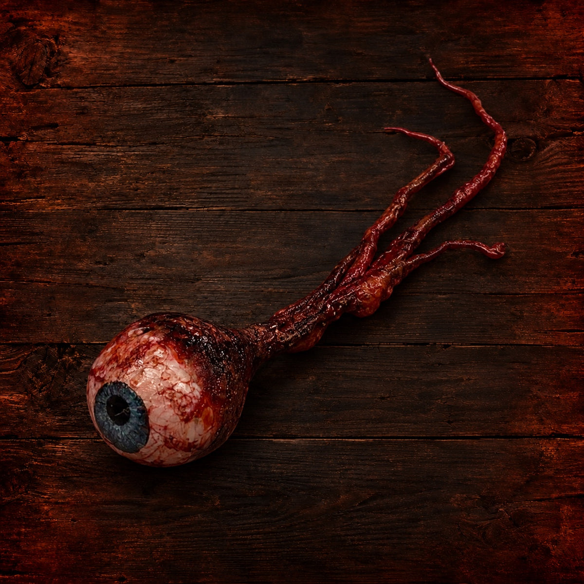 Handmade gory severed eyeball prop with a blue iris and attached optic nerve on dark wood